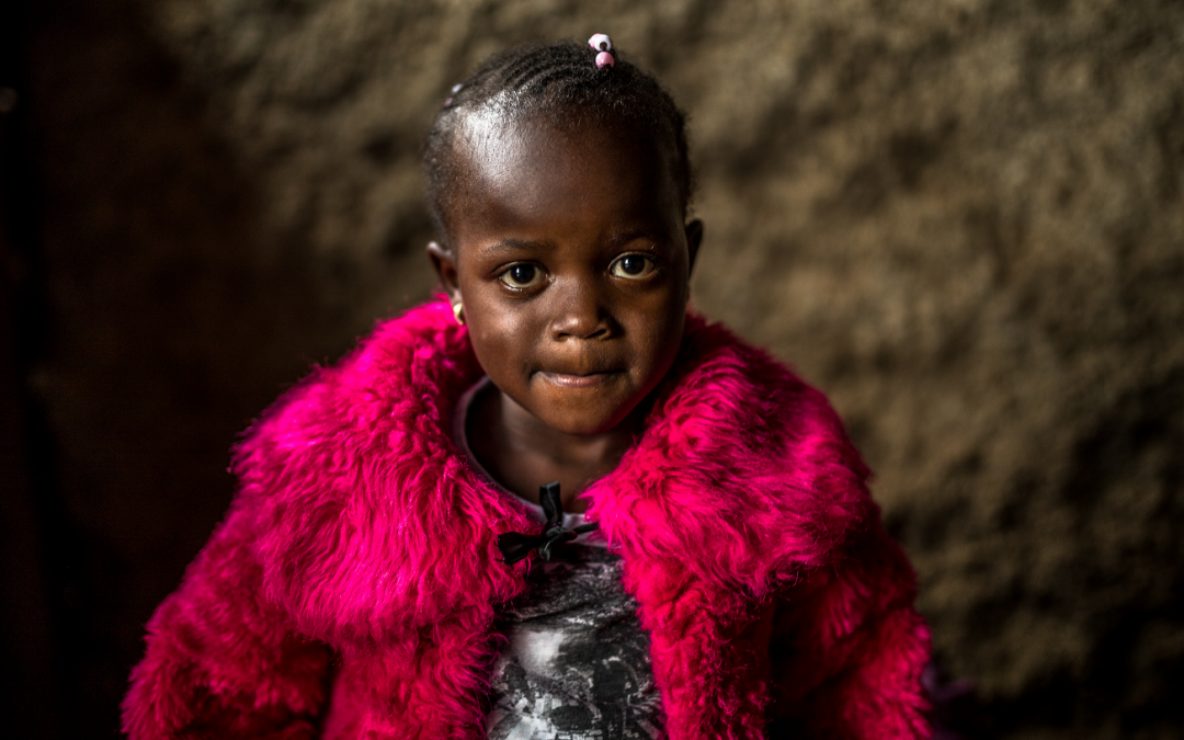 The photo shows Shazline, a four-year-old Kenyan girl with braided hair, looking into the camera, wearing a bright pink jacket.